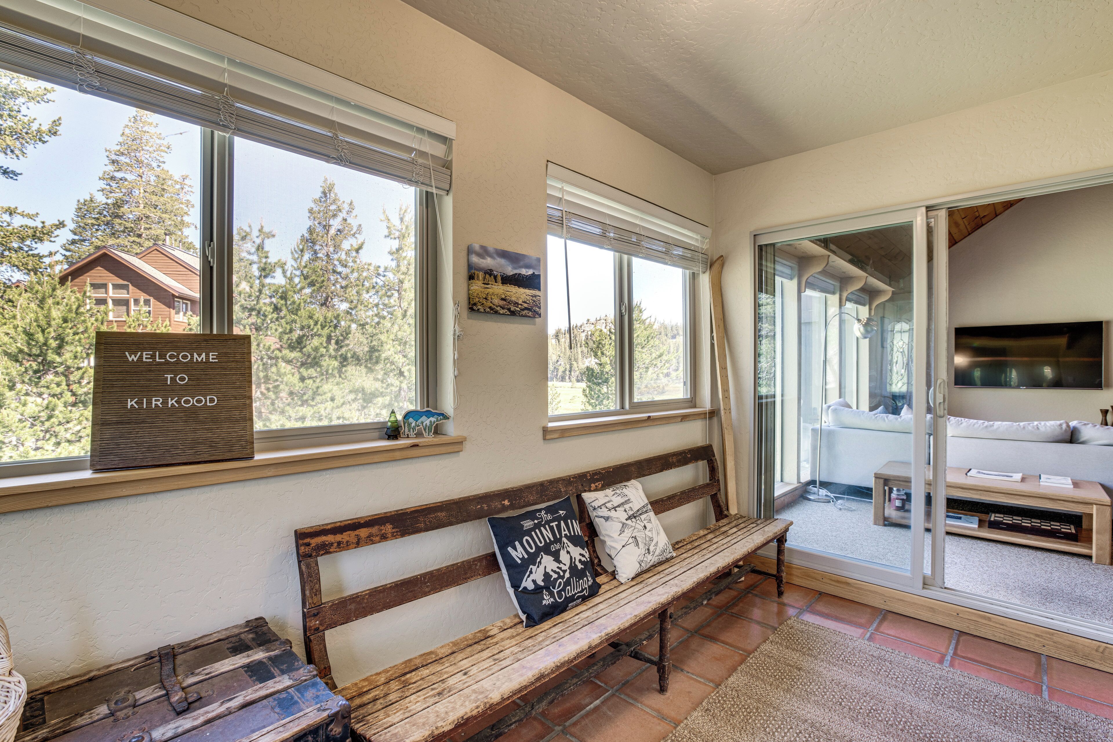 A wooden bench with a welcome sign, a view of trees and a house through large windows, and a sliding glass door leading to a living area.