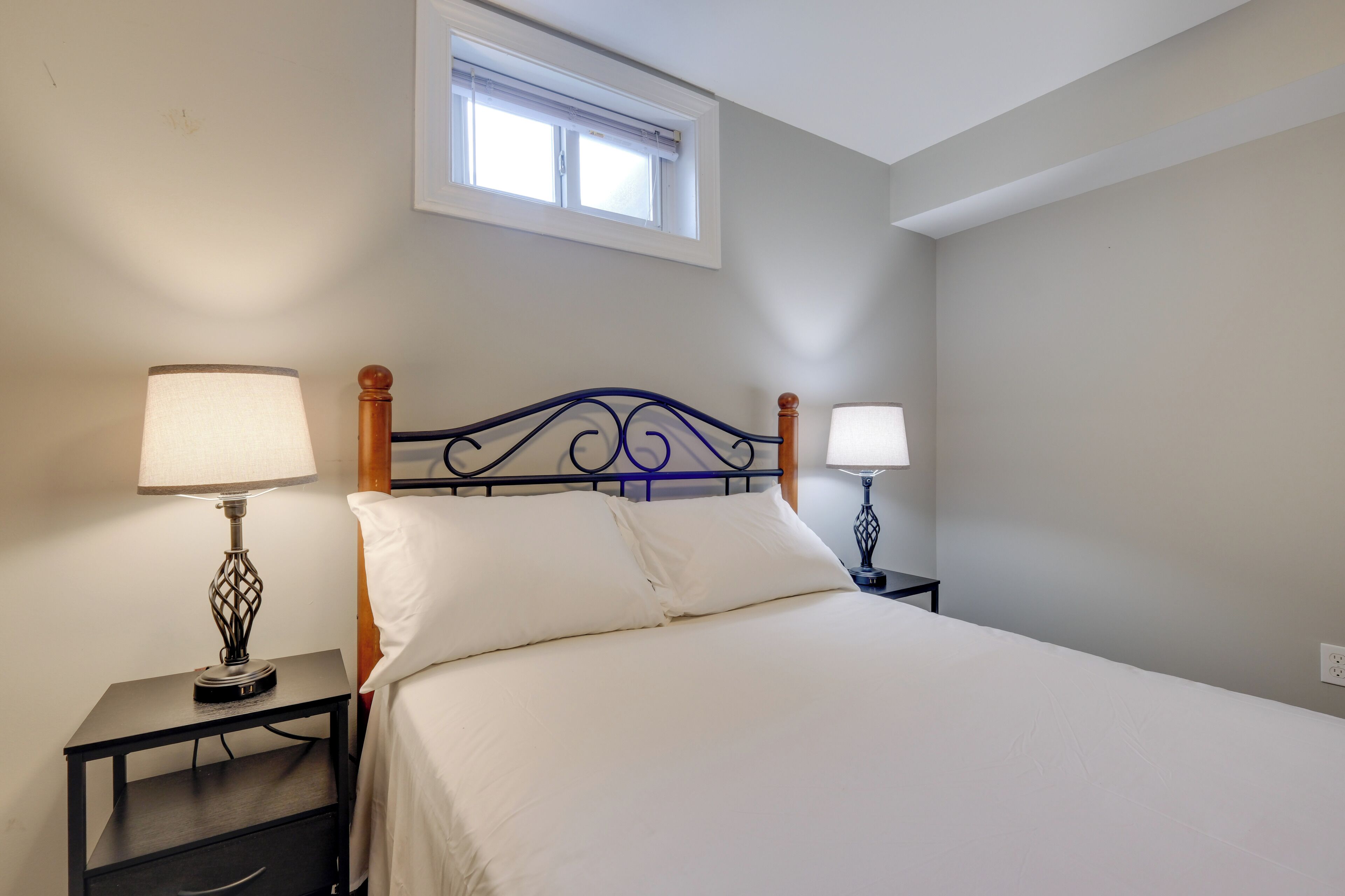A neatly made bed with white linens, two bedside lamps, a small nightstand, and a window above the bed.