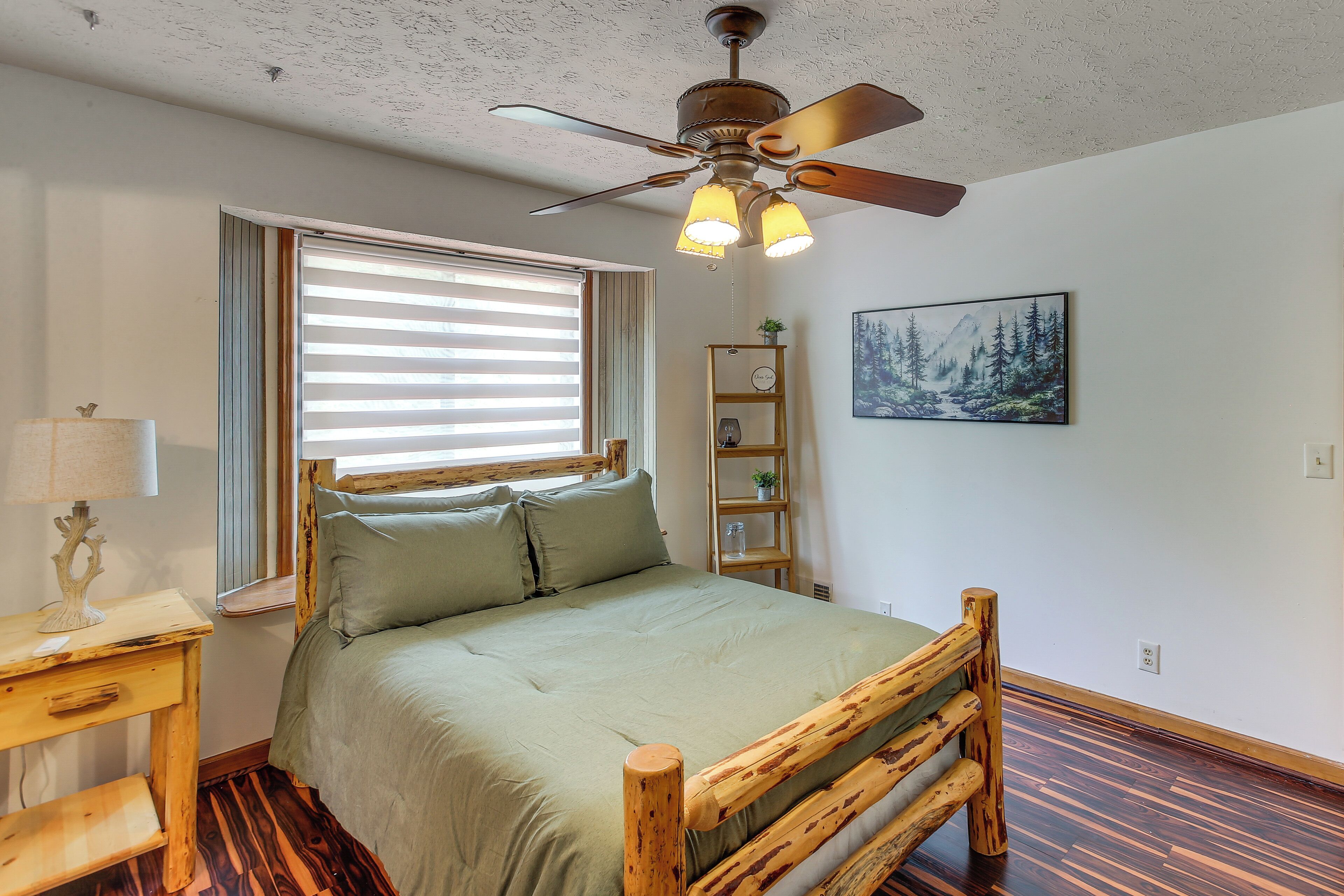 A bedroom with a bed, wooden headboard, a ceiling fan, a window with blinds, a bedside table with a lamp, a wall-mounted shelf with a clock, and a painting.