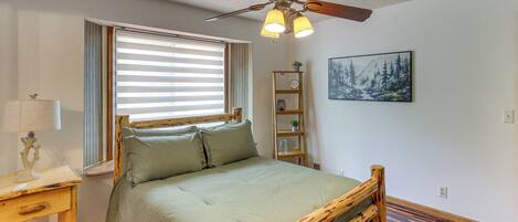 A bedroom with a bed, wooden headboard, a ceiling fan, a window with blinds, a bedside table with a lamp, a wall-mounted shelf with a clock, and a painting.