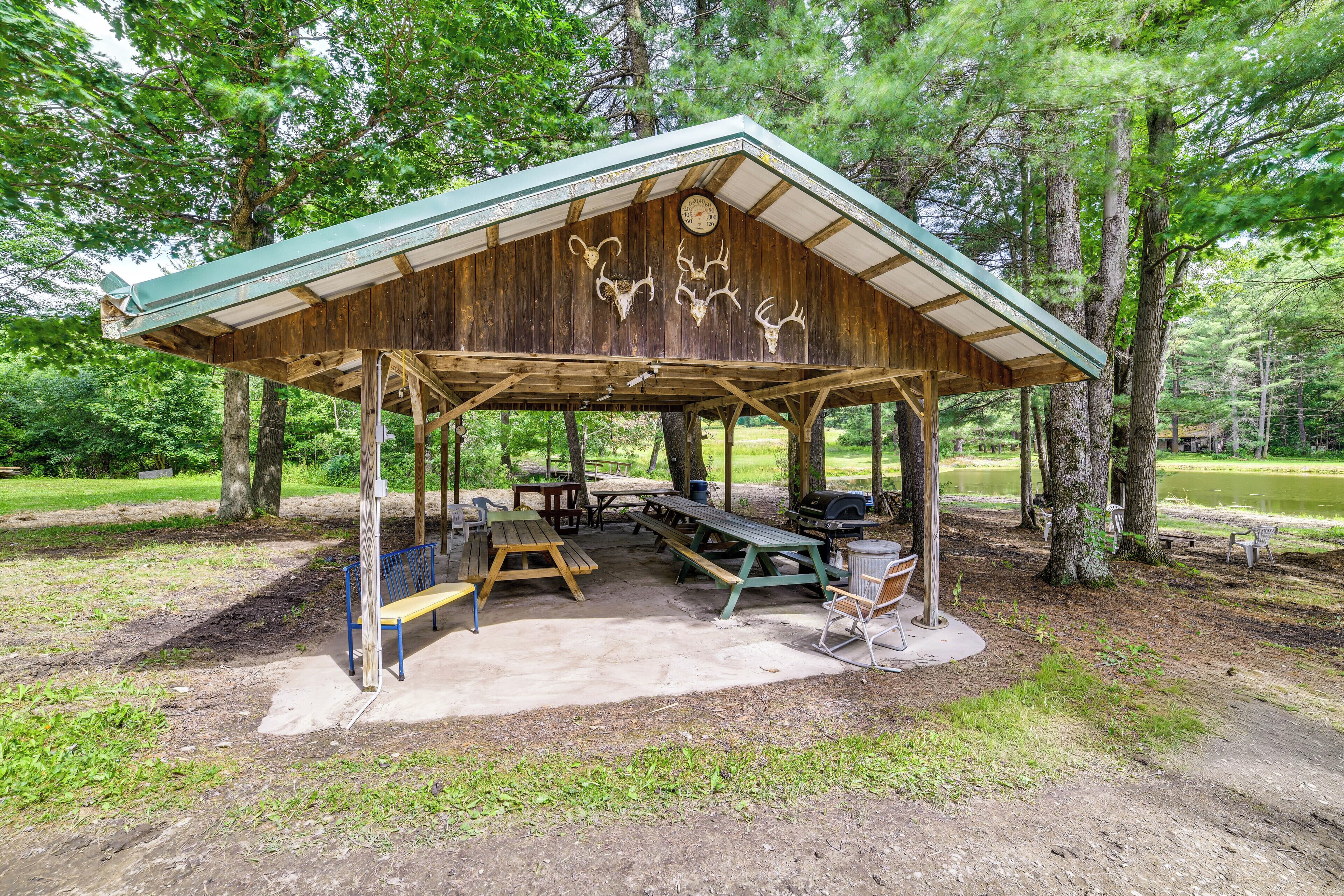 A rustic wooden pavilion with a green roof, surrounded by trees, featuring picnic tables and benches.