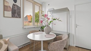 Un espace repas moderne avec une table ronde blanche, des chaises à motifs, un vase avec des fleurs et une fenêtre avec des stores.