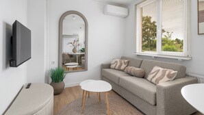 Un salon moderne avec un canapé gris, deux petites tables d’appoint blanches, une table basse ronde, une plante en pot et un téléviseur fixé au mur.