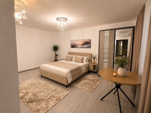 Ein modernes Schlafzimmer mit einem Bett, Nachttischen, einem runden Tisch mit einer Vase, einer Topfpflanze und einem Bild an der Wand.