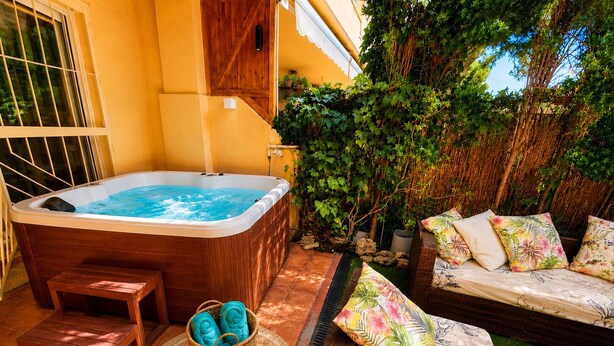 Ett bubbelbad, en träterrass, en korg av rotting med handdukar och en soffa med blommiga kuddar.