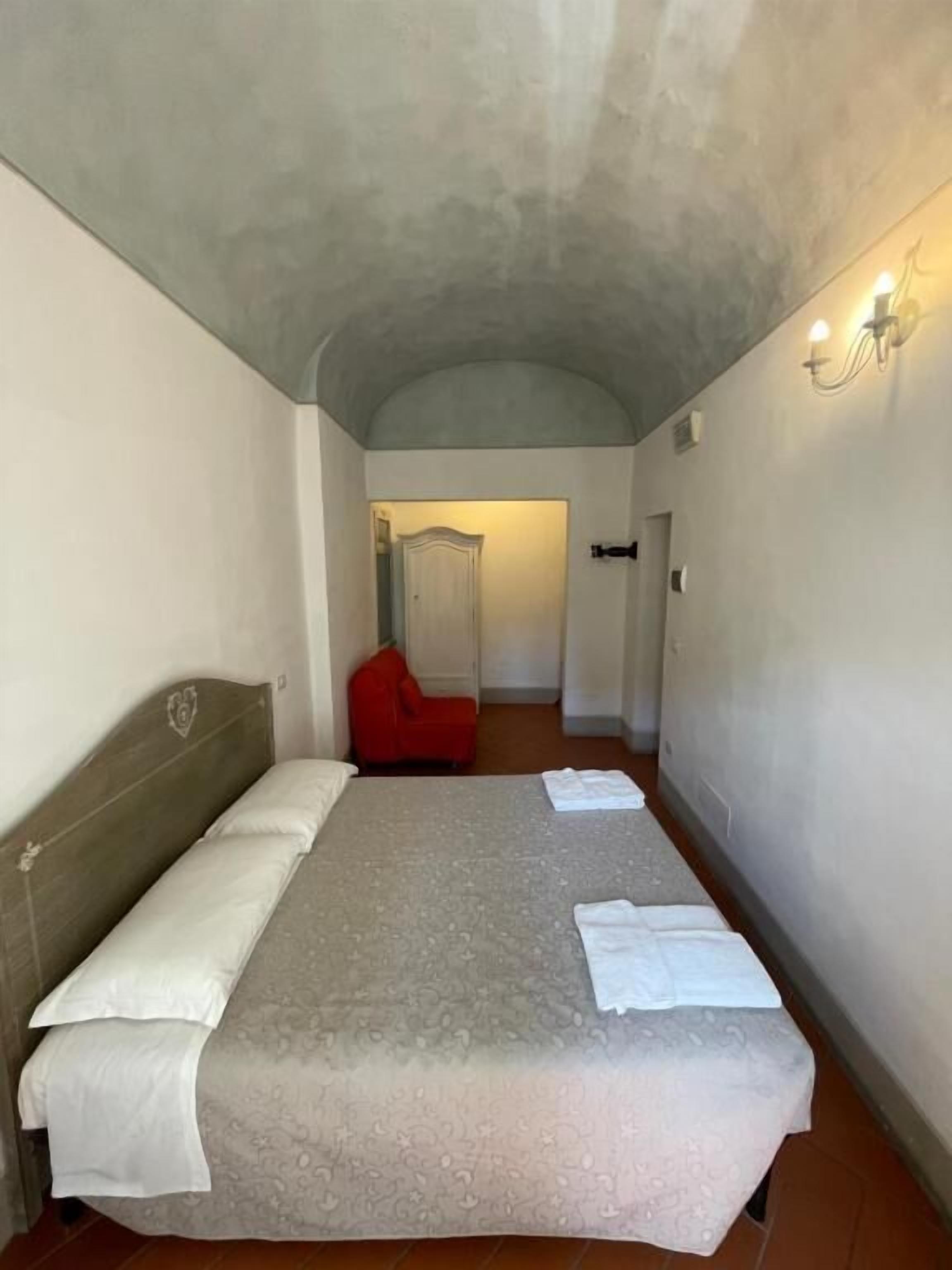 A room with a bed, a red chair, a wall-mounted light fixture, and a door leading to another room.