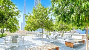 Een terras met tafels en stoelen, omringd door bomen, met uitzicht op een stadslandschap met een opvallende wolkenkrabber.