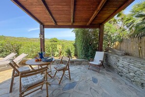 A covered patio with stone flooring, wooden furniture, and a view of a lush landscape. - Relax at Lentischi – Private Veranda & BBQ (Stintino)