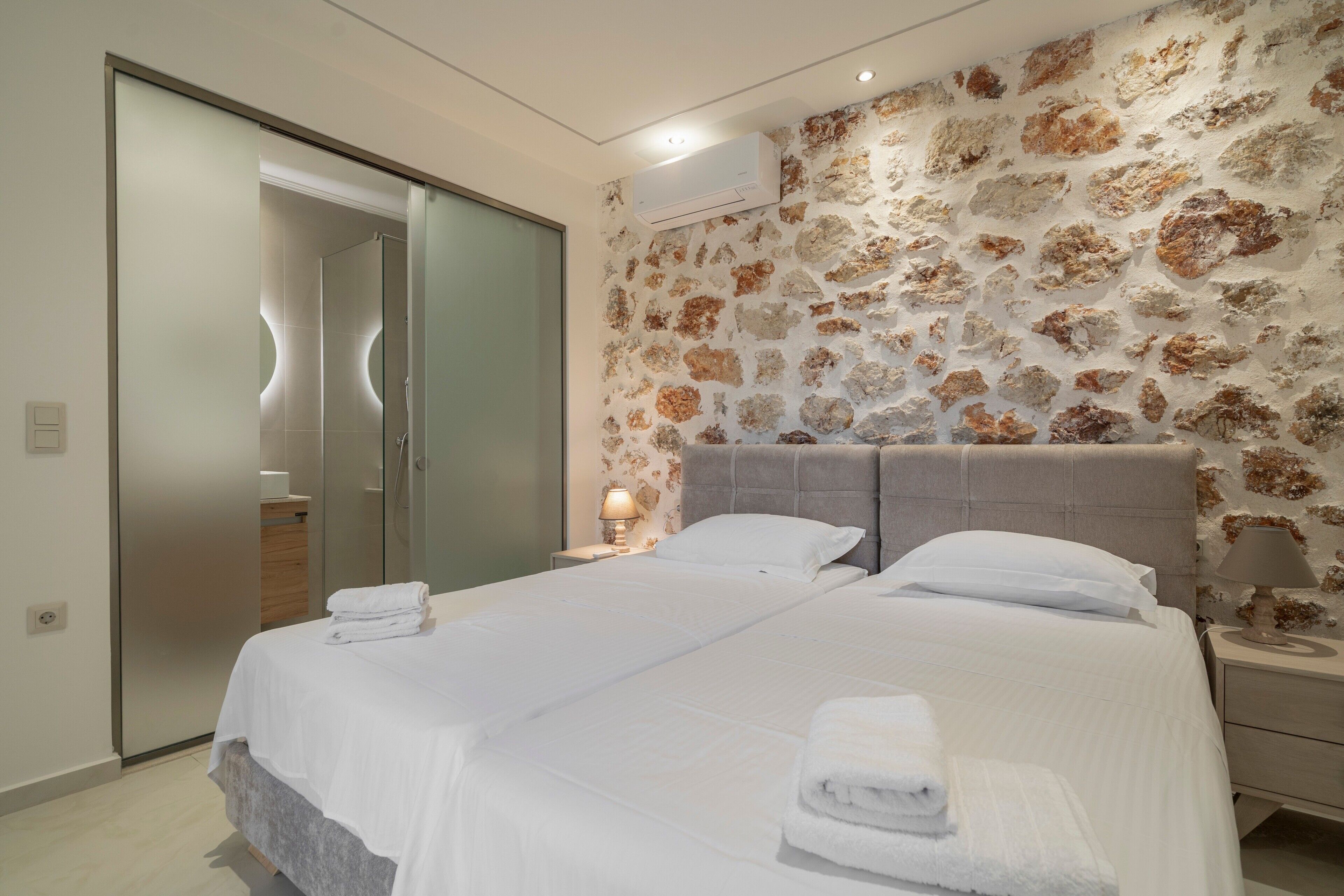 A modern bedroom with a large bed, two bedside tables, a nightstand, and a wall-mounted air conditioning unit.