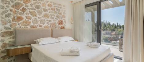 Un dormitorio con pared de piedra, cama con ropa blanca, mesita de noche y vistas a un balcón.