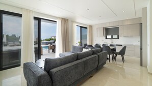 Ein modernes Wohnzimmer mit einer grauen Eckcouch, einem Esstisch mit Stühlen und einer Küchenzeile mit weißen Schränken sowie einem Flachbildfernseher.