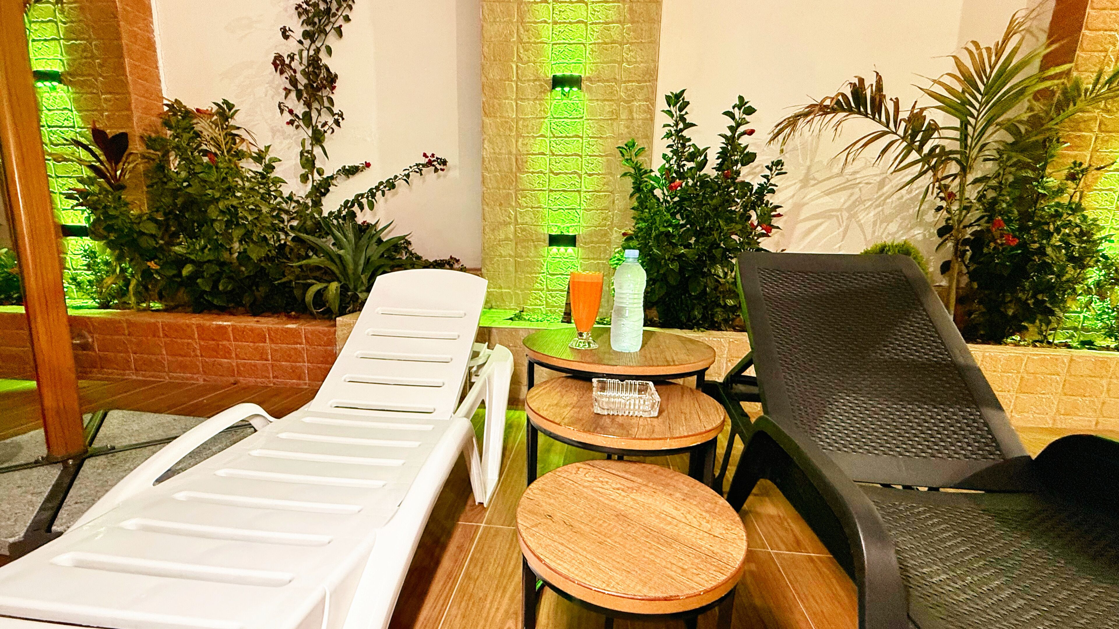 A sun lounge area with a white lounge chair, a black lounge chair, a small round table with a bottle and glass, and a brick wall with green foliage.