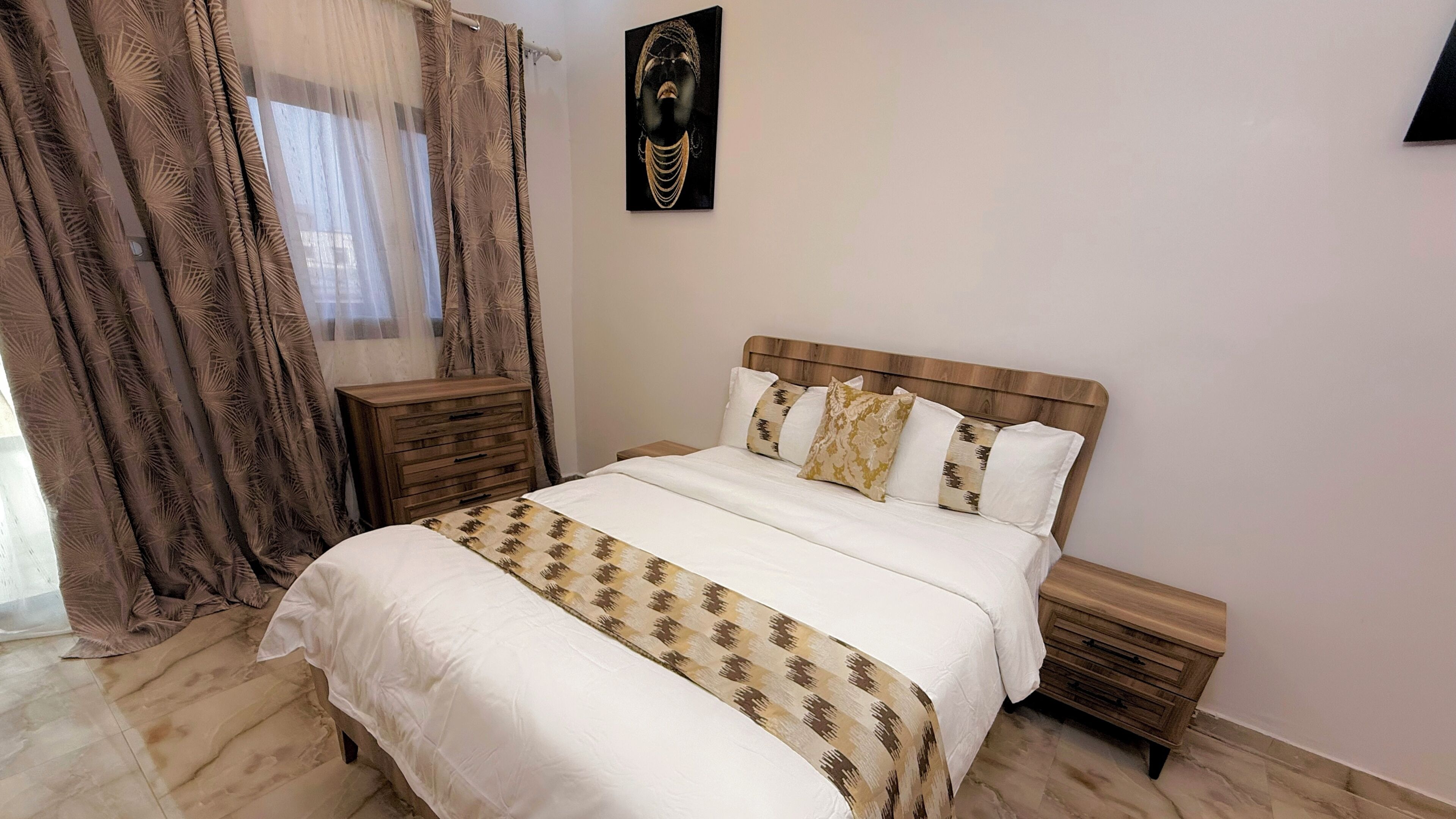 Un dormitorio con cama, mesitas de noche, cabecera de madera, una obra de arte en la pared y una ventana con cortinas.