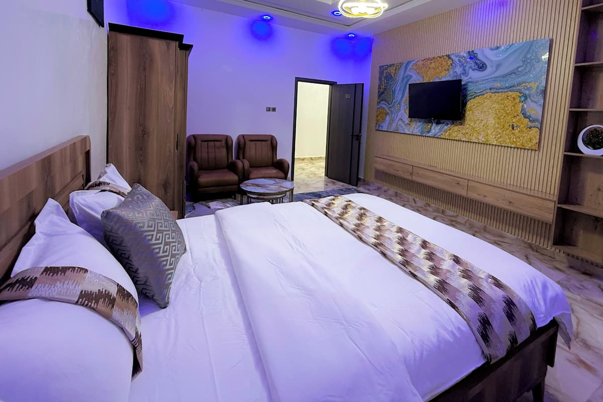 Habitación de hotel moderna con una cama grande, dos sillones, una mesita, un televisor montado en una pared de madera y una puerta que da a otra habitación.