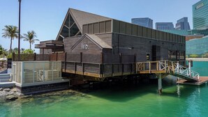 Un edificio moderno con fachada de madera y cristal, ubicado junto a una costa con un muelle y una escalera que lo conecta.