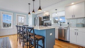 Een moderne keuken met een centraal eiland, krukken, roestvrijstalen apparatuur en hanglampen.