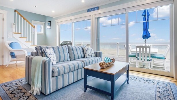 Een woonkamer met een gestreepte bank, een houten bijzettafeltje, een rieten mand, een blauwe paraplu en uitzicht op het strand.