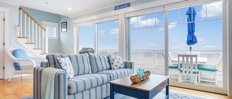A living room with a striped sofa, a wooden coffee table, a wicker basket, a blue umbrella, and a view of the beach.