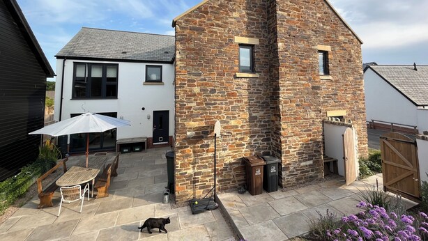 Een stenen gebouw met schoorsteen, een terras met tafel en stoelen, en een kat die over het terras loopt.