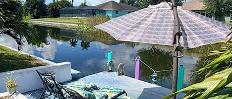A tranquil outdoor seating area with a table and chairs, a covered umbrella, and a calm water body reflecting the sky.