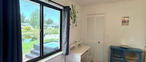 A bedroom with a bed, a window with curtains, a bedside table, a wardrobe, and a view of a backyard with a pool.