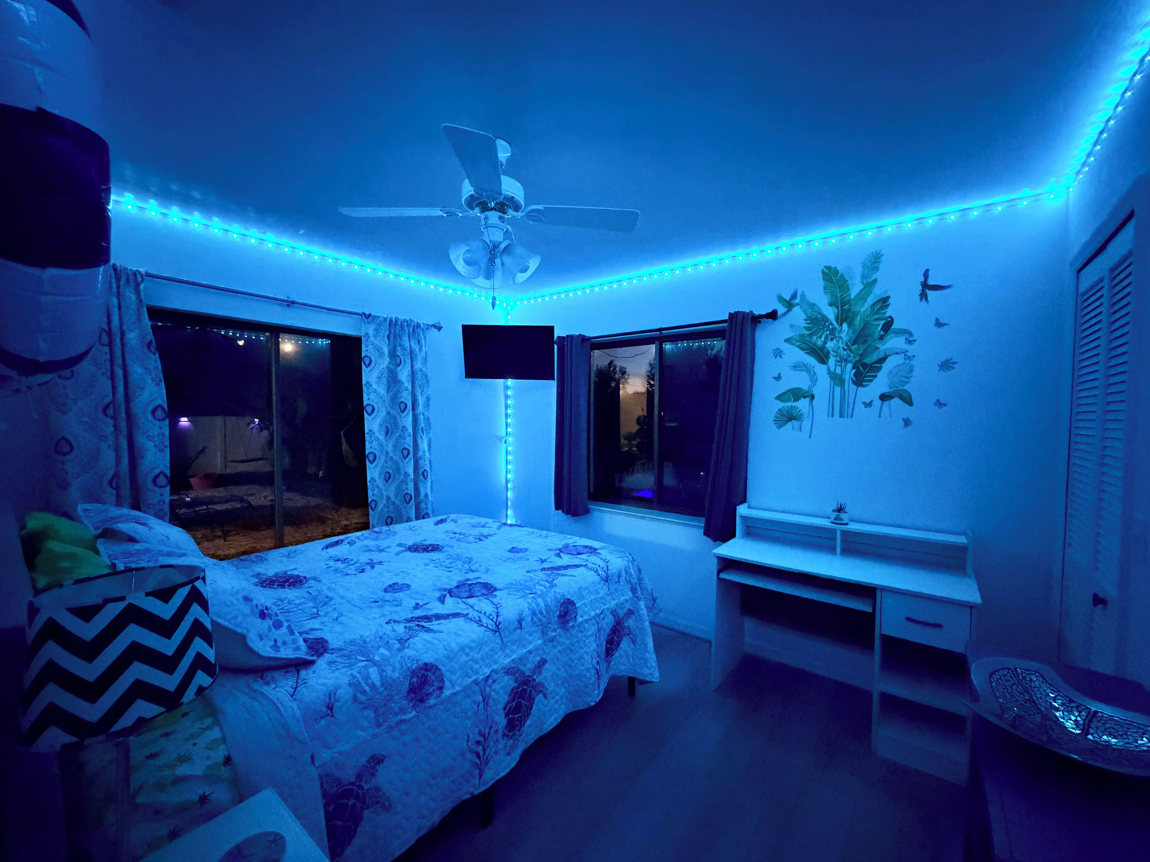 Un dormitorio con cama, ventilador de techo, ventana y pared con un adhesivo de planta.
