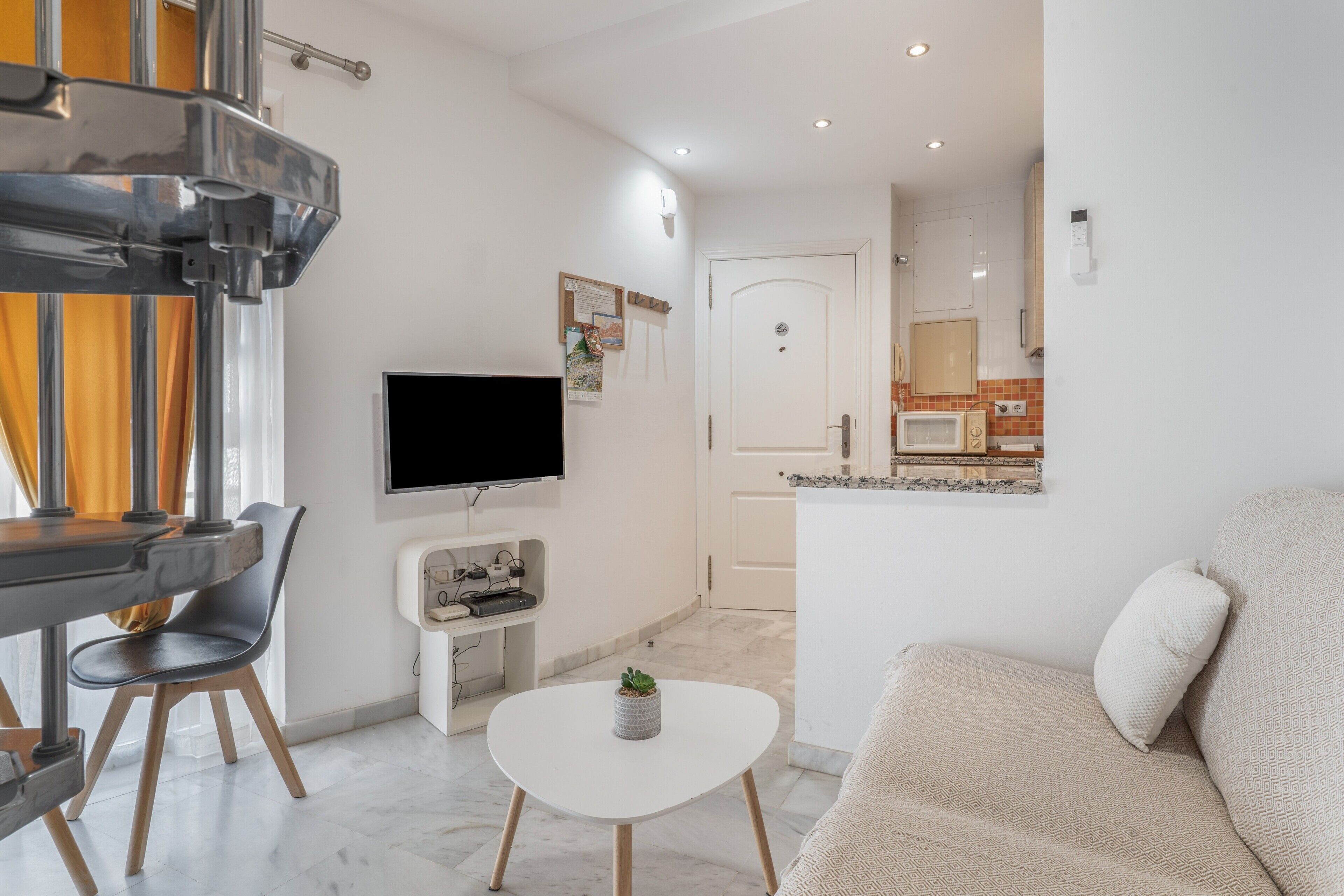 A modern living room with a flat-screen TV, a small table with a plant, a sofa, and a kitchen area in the background.