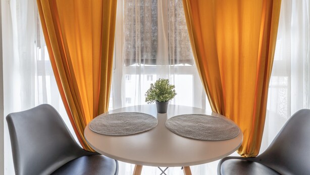 Une table ronde avec deux chaises, une petite plante en pot et une fenêtre ornée de rideaux légers.