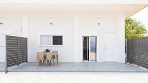 A modern building with a covered patio area featuring a table and chairs, a window, and a door. - Cozy Coastal Apartment in Oropos! Perfect for Relaxing Getaways! (Νέα Παλάτια)