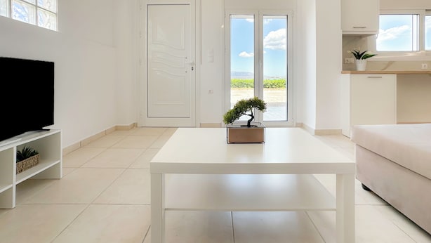 A modern living room with a flat-screen TV, a white coffee table, and a sliding glass door leading to a balcony with a view of the countryside. - Cozy Coastal Apartment in Oropos! Perfect for Relaxing Getaways! (Νέα Παλάτια)