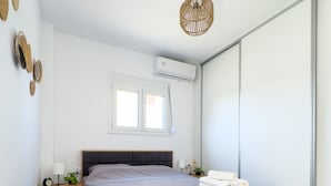 A modern bedroom with a wooden bed frame, a grey bedspread, a bedside table with a lamp, a wall-mounted air conditioner, and a window with natural light. - Cozy Coastal Apartment in Oropos! Perfect for Relaxing Getaways! (Νέα Παλάτια)