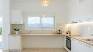 A modern kitchen with white cabinetry, a wooden countertop, a built-in oven, and a window with a mountain view. - Cozy Coastal Apartment in Oropos! Perfect for Relaxing Getaways! (Νέα Παλάτια)