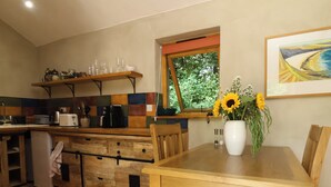 A kitchen with wooden cabinets, a dining table with chairs, a window with a view of greenery, and a vase with flowers on the table. - Brambly Cabin (Crediton)