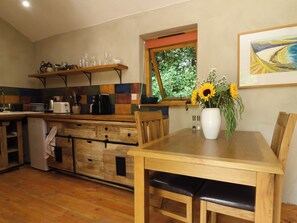 Una cocina con armarios de madera, una mesa de comedor con sillas, un ventanal con vistas a la vegetación y un jarrón con flores sobre la mesa.