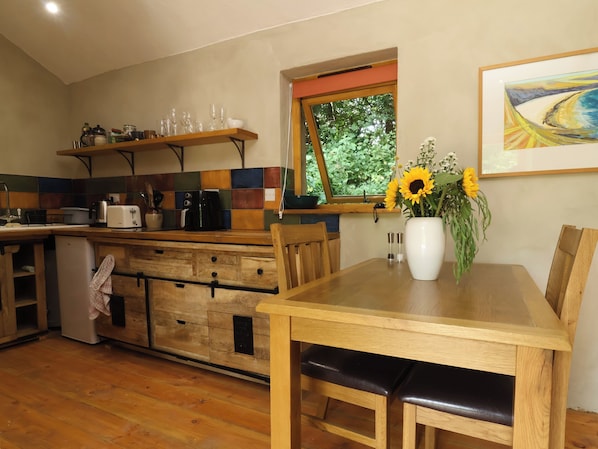Una cocina con armarios de madera, una mesa de comedor con sillas, un ventanal con vistas a la vegetación y un jarrón con flores sobre la mesa.
