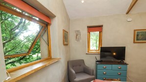 A bedroom with a bed, a television on a dresser, a chair, and a window with a view of trees. - Brambly Cabin (Crediton)