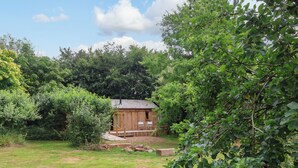 A wooden cabin with a porch, surrounded by a grassy yard and dense foliage. - Brambly Cabin (Crediton)