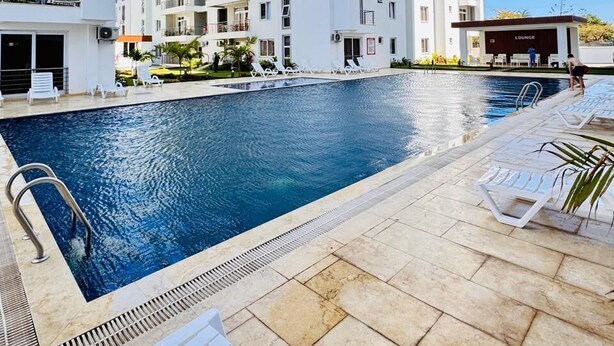 Piscina com espreguiçadeiras, um edifício com varandas e um céu azul límpido.