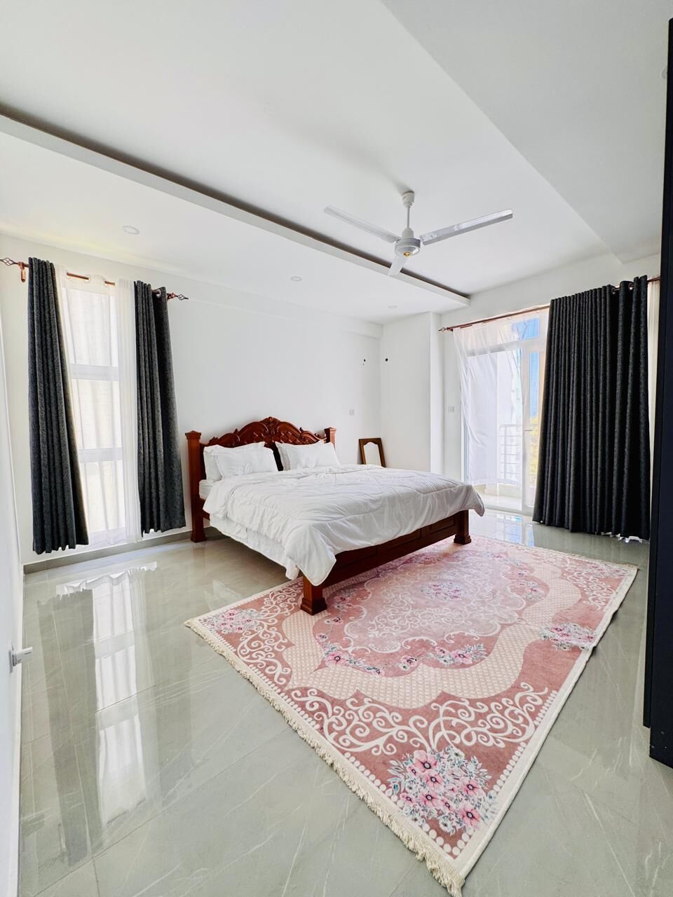 A bedroom with a bed, a ceiling fan, a large window, and a patterned rug.