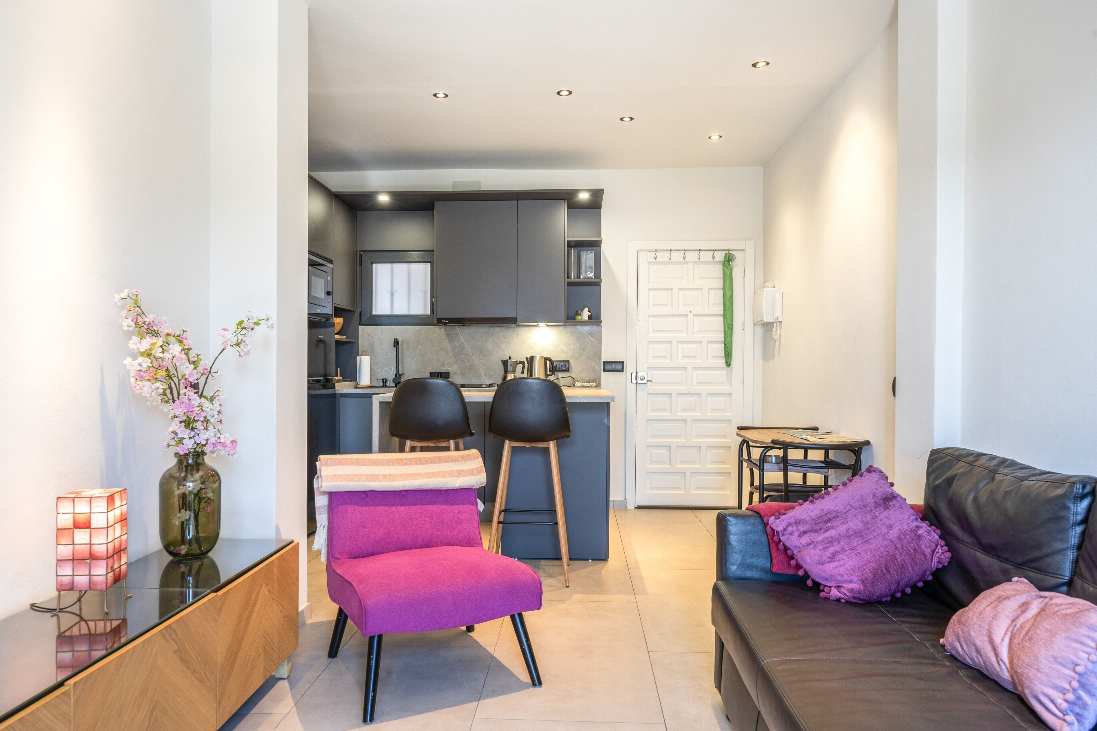 A modern living room with a sofa, chairs, a small table, and a kitchen in the background.