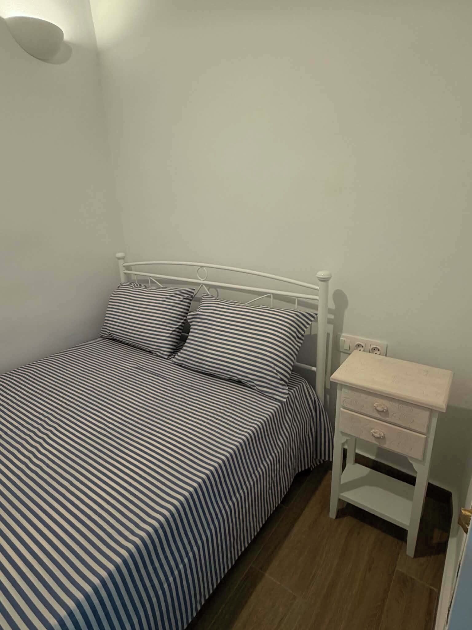 Enkeltseng med sengetæppe med striber, et natbord med skuffer og en vægmonteret lampe.