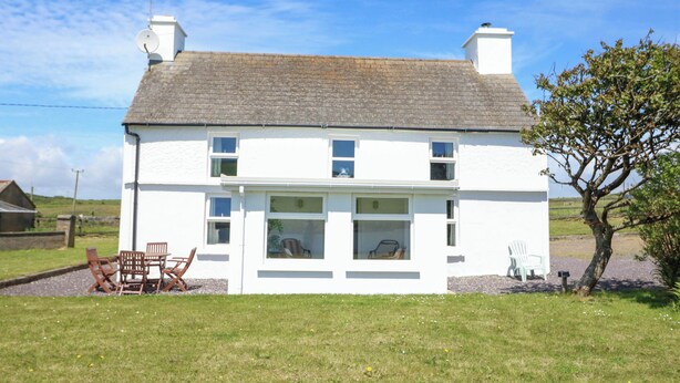 A two-story white house with a grey roof, a chimney, and a small garden with a tree and a chair. - Teach Feirme (Clonakilty)