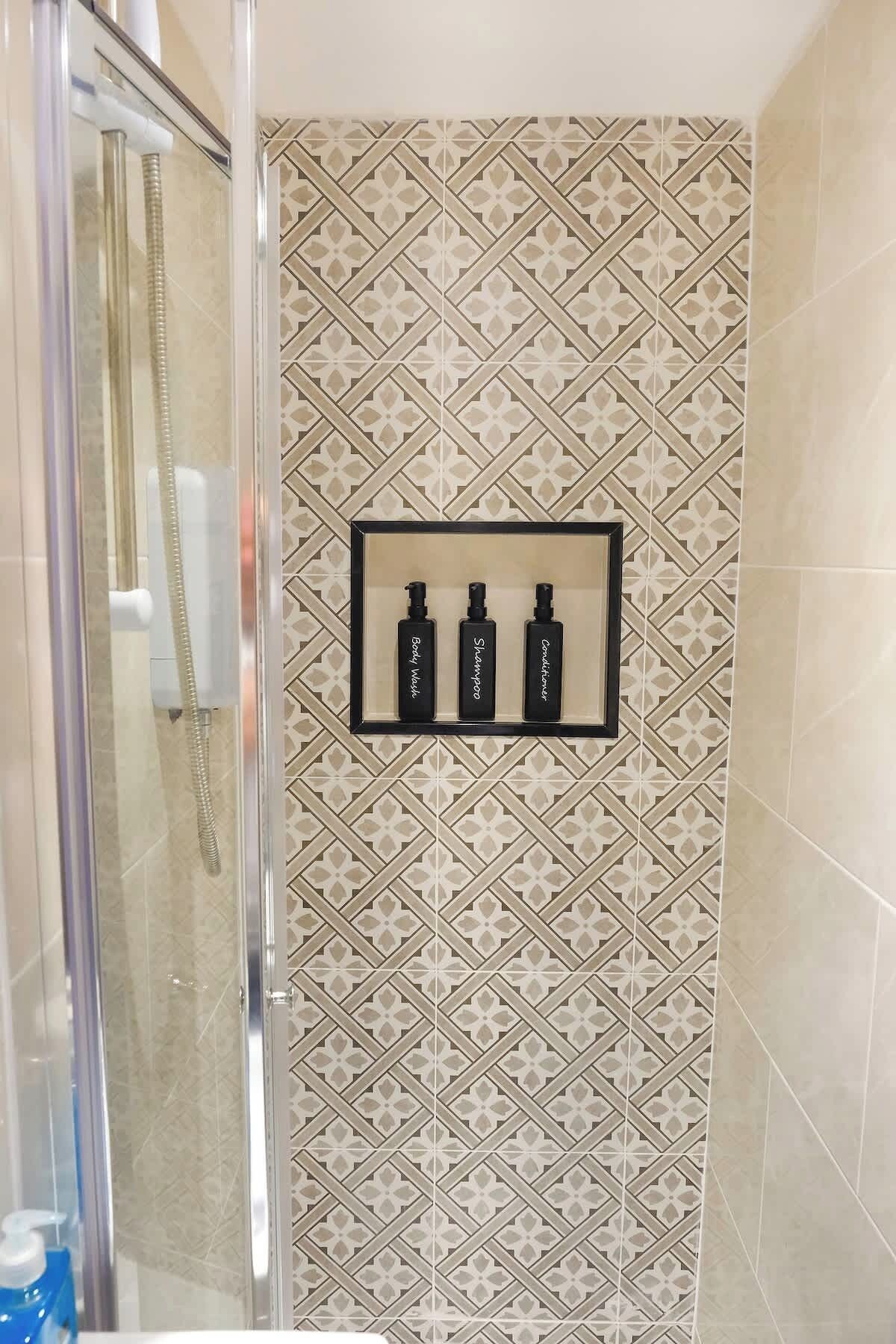Une douche avec une porte en verre, un mur recouvert de carreaux décoratifs et une étagère sur laquelle sont posées trois bouteilles étiquetées « gel douche », « shampoing » et « après-shampoing ».