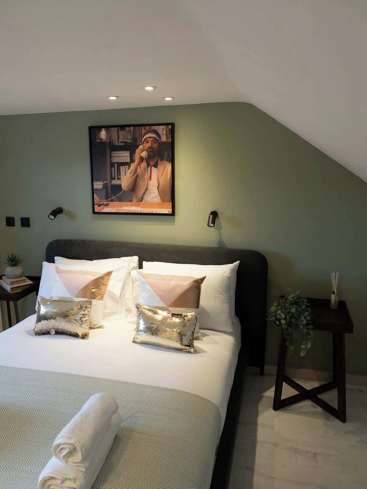 A bedroom with a bed, pillows, a painting, a small table with a plant, and a wall-mounted light.