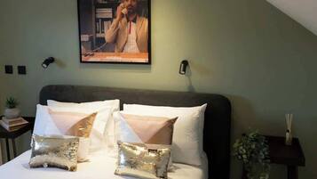 A bedroom with a bed, pillows, a painting, a small table with a plant, and a wall-mounted light.