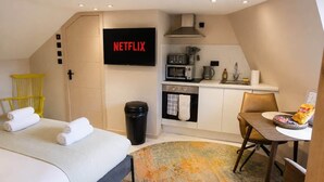 A hotel room with a bed, a desk with a chair, a kitchenette, a TV, and a trash bin.
