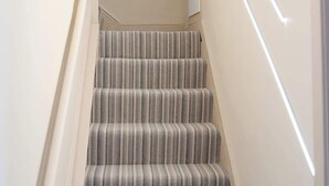 Un escalier recouvert d’une moquette à rayures, avec une main courante et un luminaire fixé au mur.