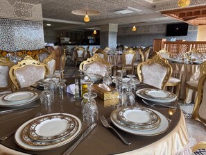 Un comedor con sillas ornamentadas en dorado y blanco, una mesa redonda dispuesta para una comida, y un televisor montado en la pared.