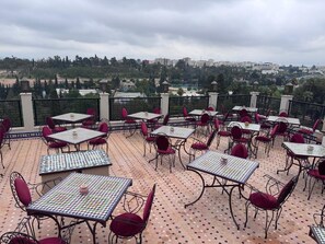 An outdoor patio area with multiple tables and chairs, a patterned floor, and a view of a cityscape in the background.