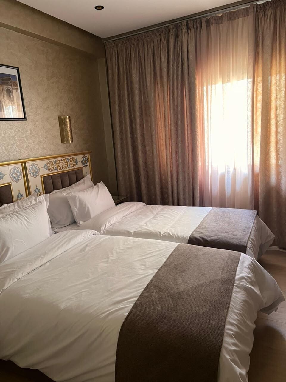 A hotel room with two beds, a headboard with decorative panels, a framed picture on the wall, and a window with curtains.